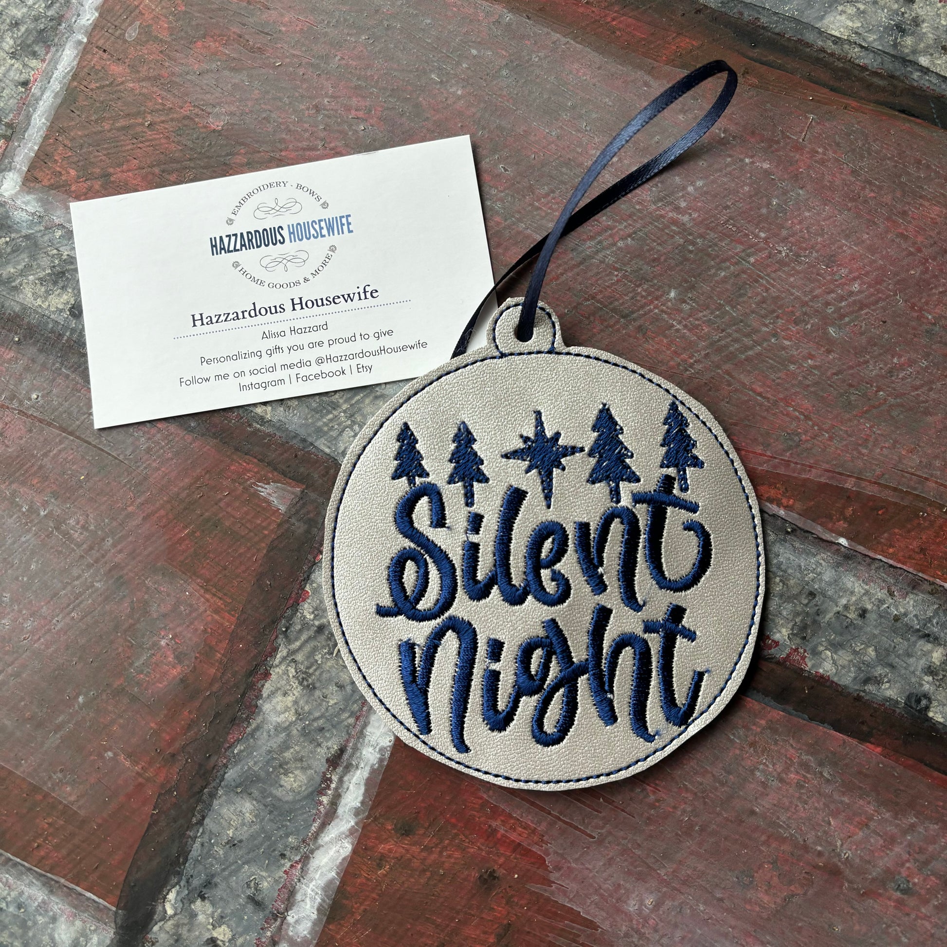 Round embroidered patch with 'Silent Night' text and trees, attached to a card on a stone surface.