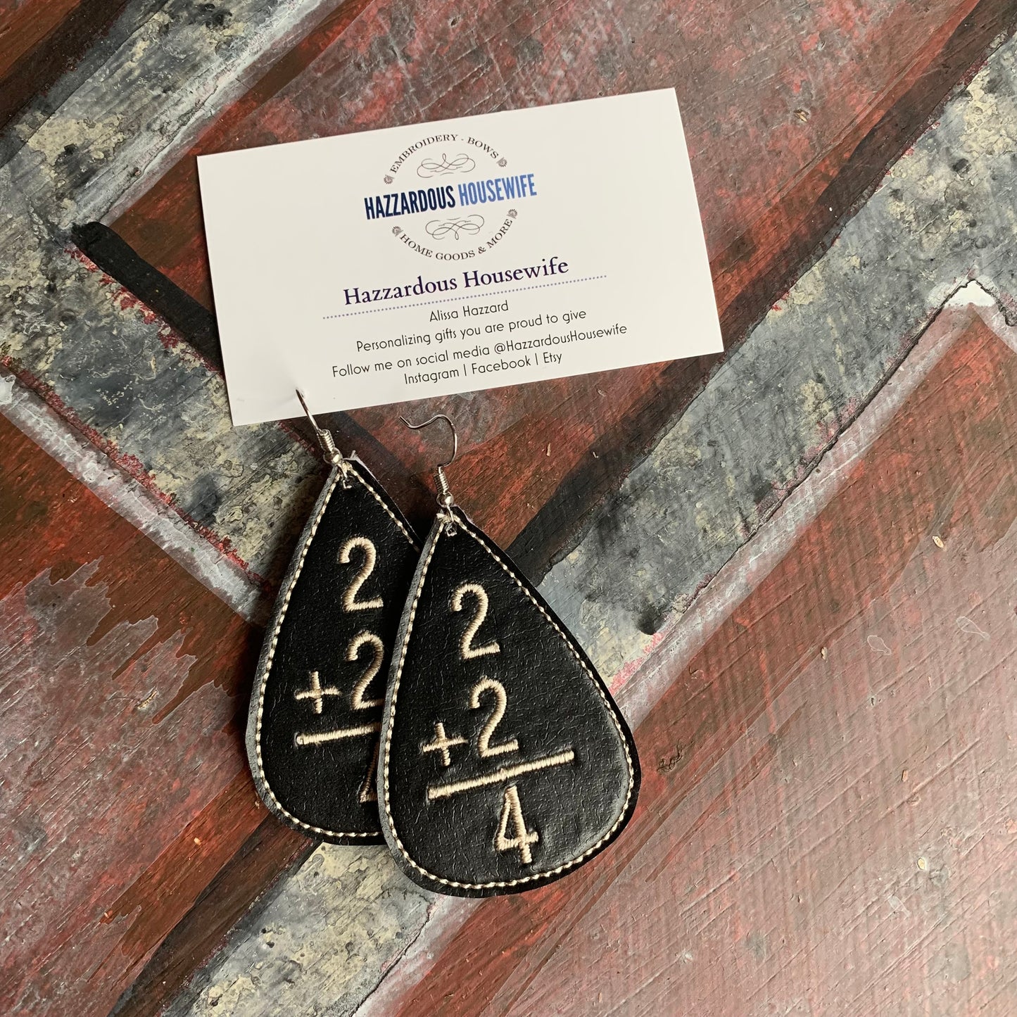 Teardrop-shaped earrings with arithmetic design on a rustic wooden surface, accompanied by a business card for 'Hazzardous Housewife'.