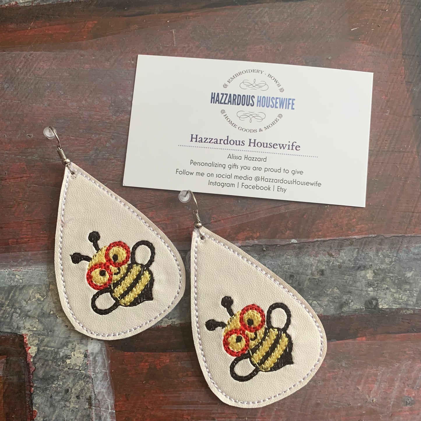 White Teardrop-shaped earrings with bee design on a wooden surface with a business card for 'Hazzardous Housewife'.