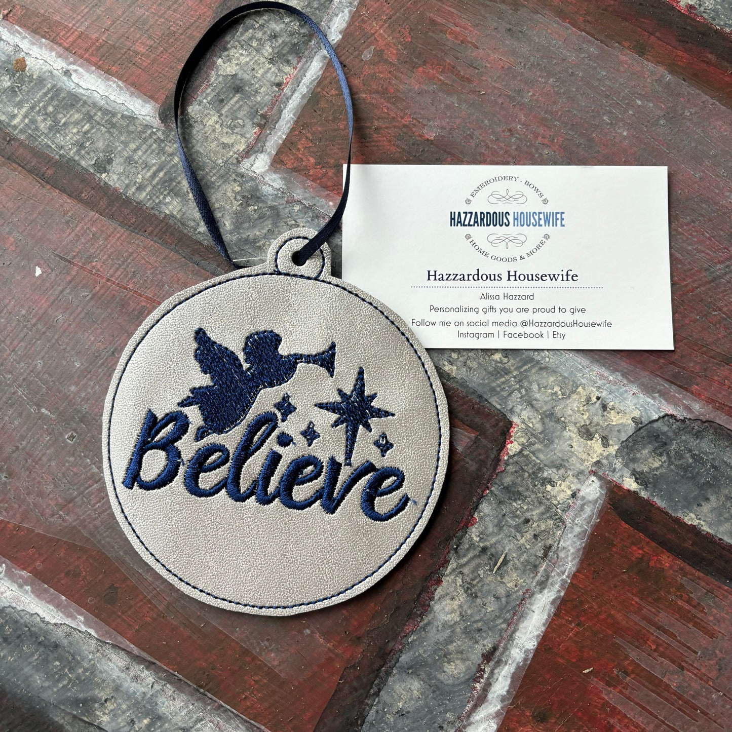 Round embroidered patch with 'Believe' text and angel design on a wooden surface, with a card labeled 'Hazzardous Housewife' in the background.