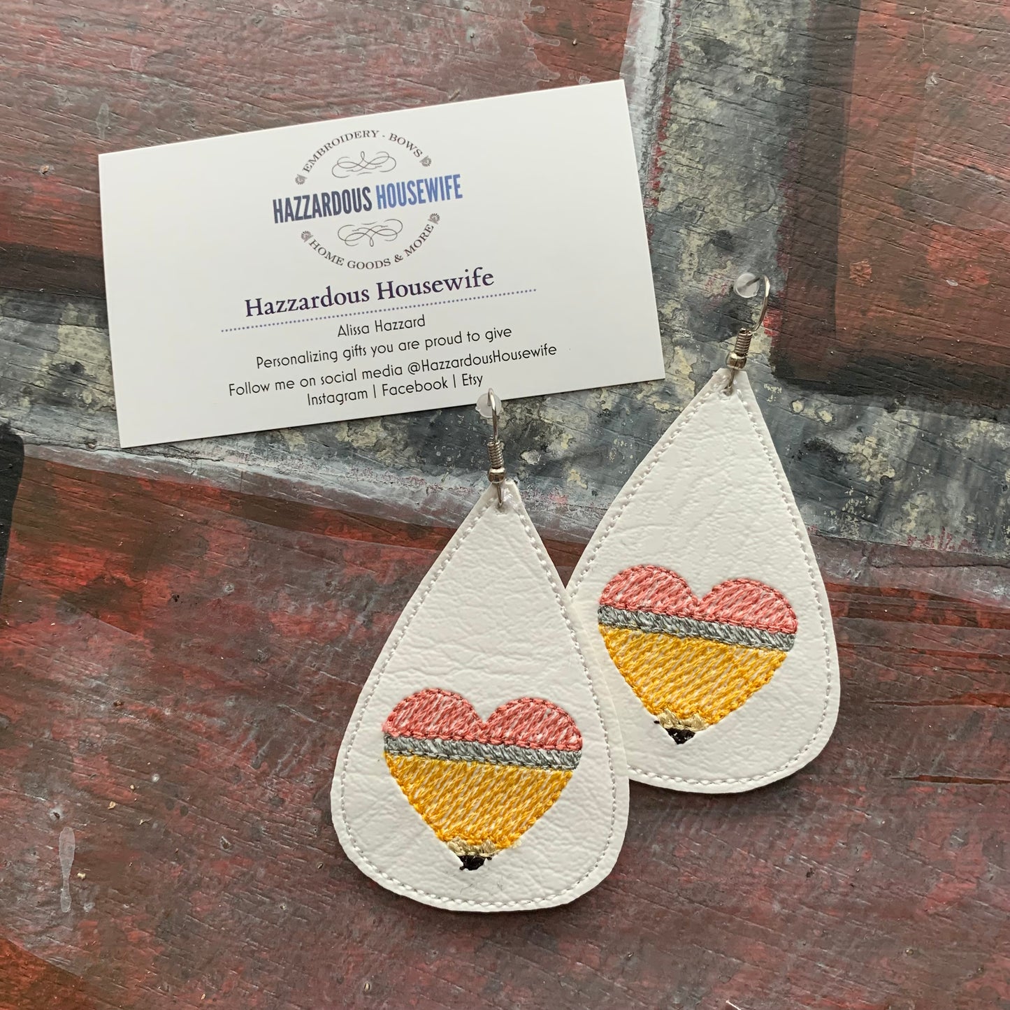 Teardrop-shaped earrings with embroidered pencil in the shape of a heart design on a brick surface with a business card.