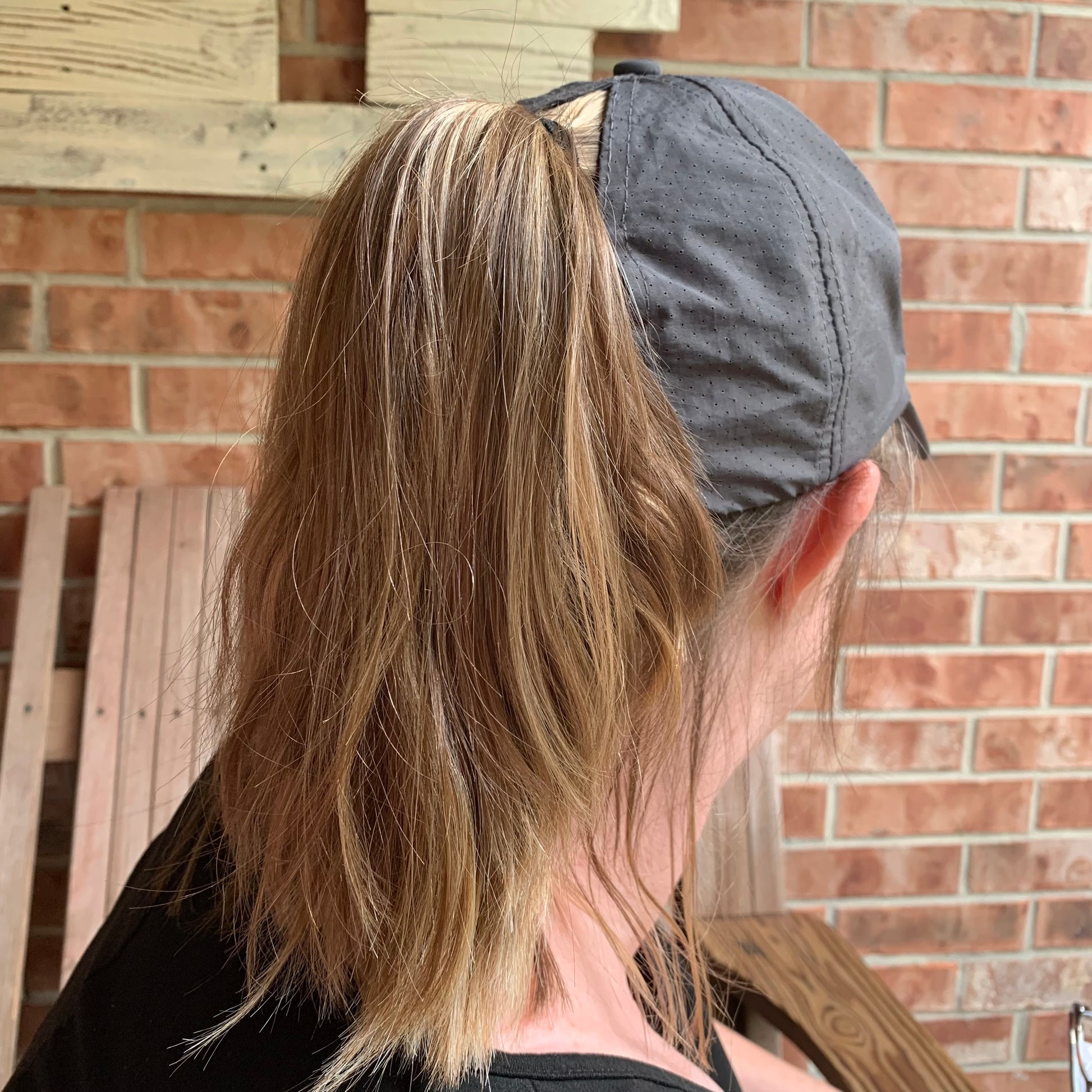 Person wearing a gray cap with long hair tied back through hole in cap, standing against a brick wall.