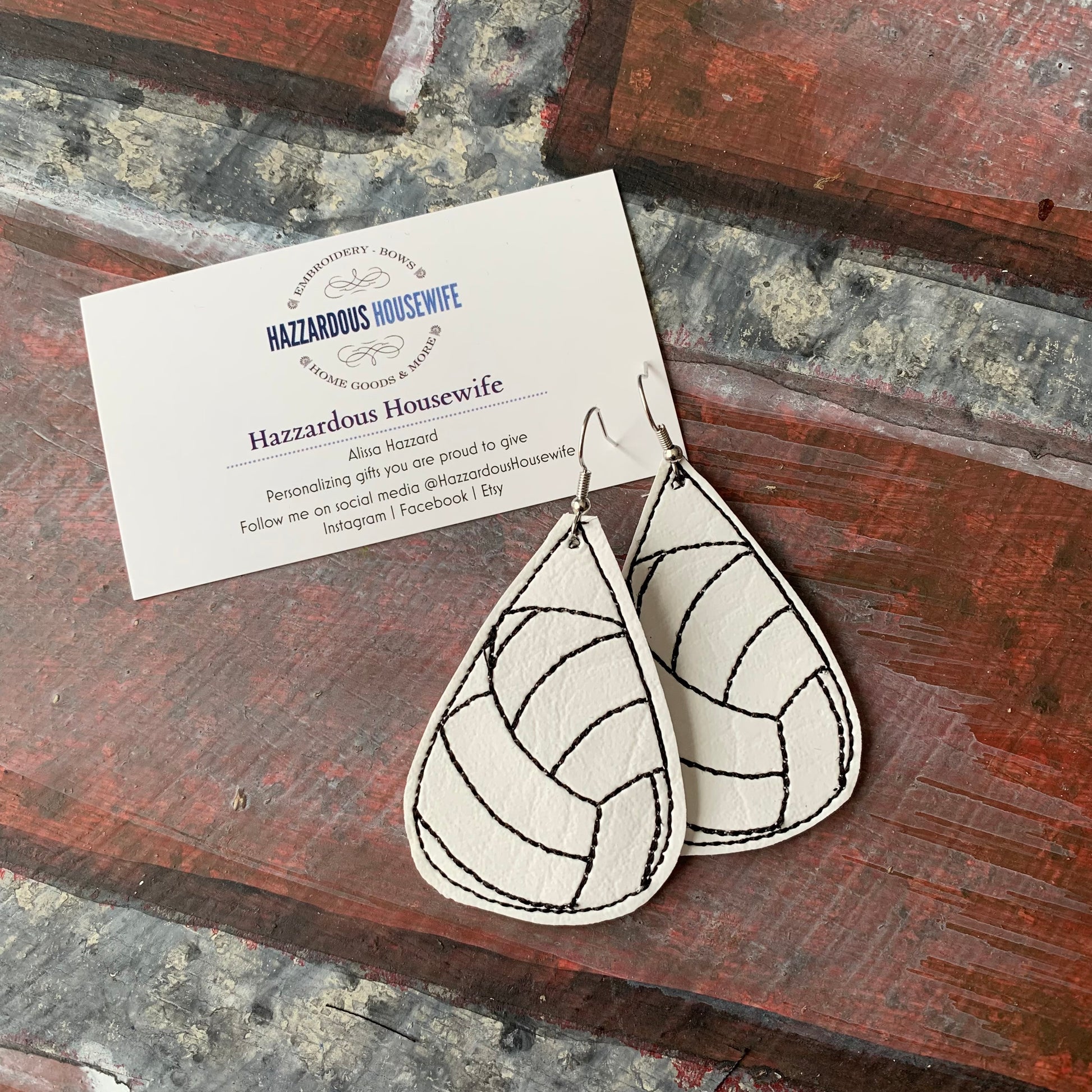 Leather volleyball earrings with a pattern on a wooden surface with a HazzardousHousewife business card.