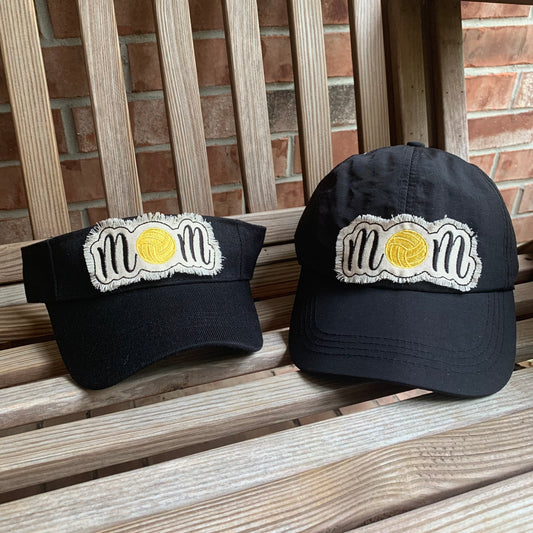Two black caps with 'mom' embroidery on a wooden surface with a brick wall background.  The 'o' is a water polo ball. 