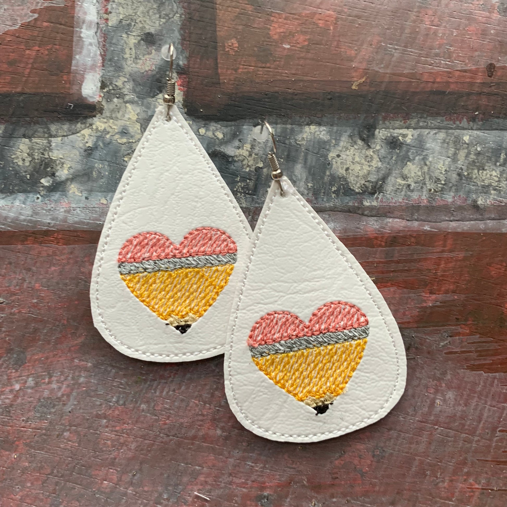 Teardrop-shaped earrings with embroidered pencil in the shape of a heart design on a brick surface.