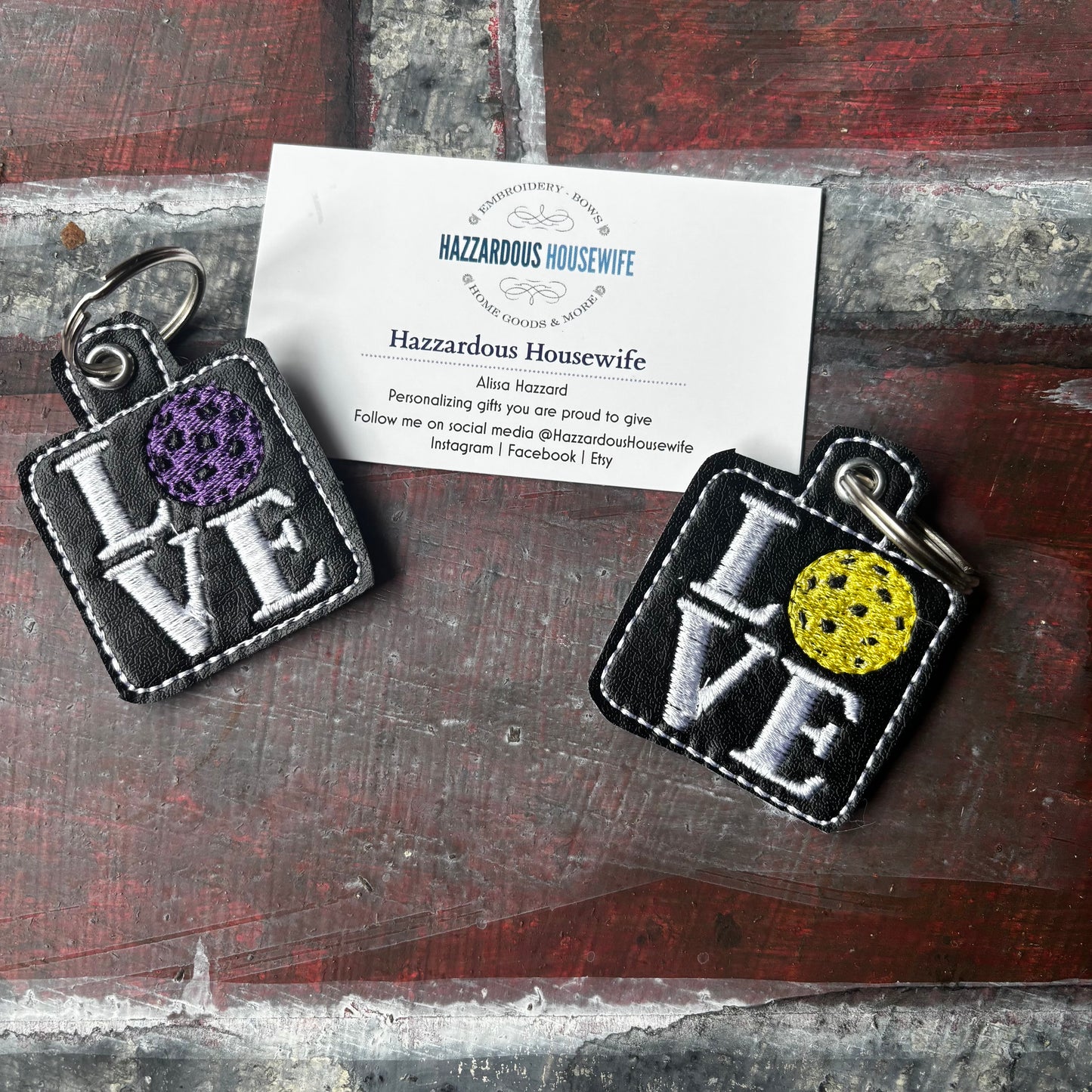 Two 'LOVE' pickleball keychains with purple and yellow accents on a rustic wooden surface with a business card.