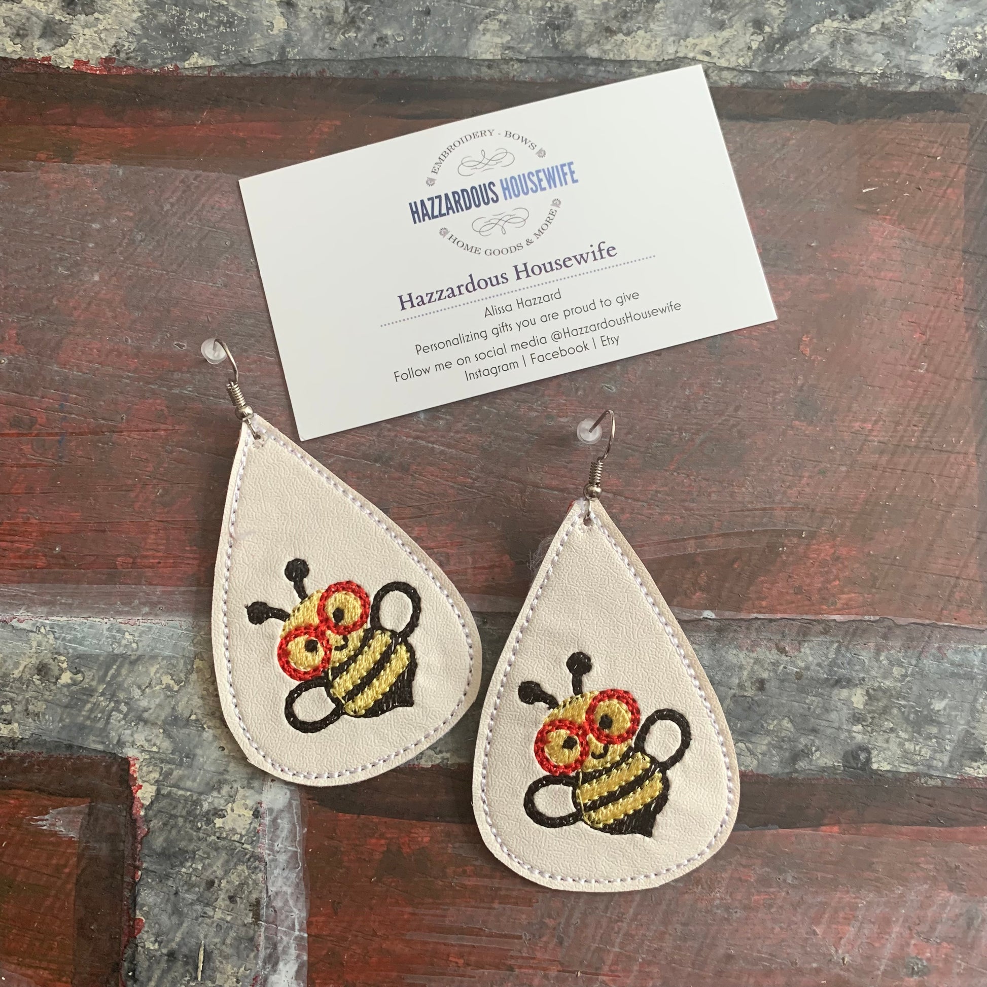 Teardrop-shaped earrings with embroidered bee design on a wooden surface, featuring 'Hazardous Housewife' branding.