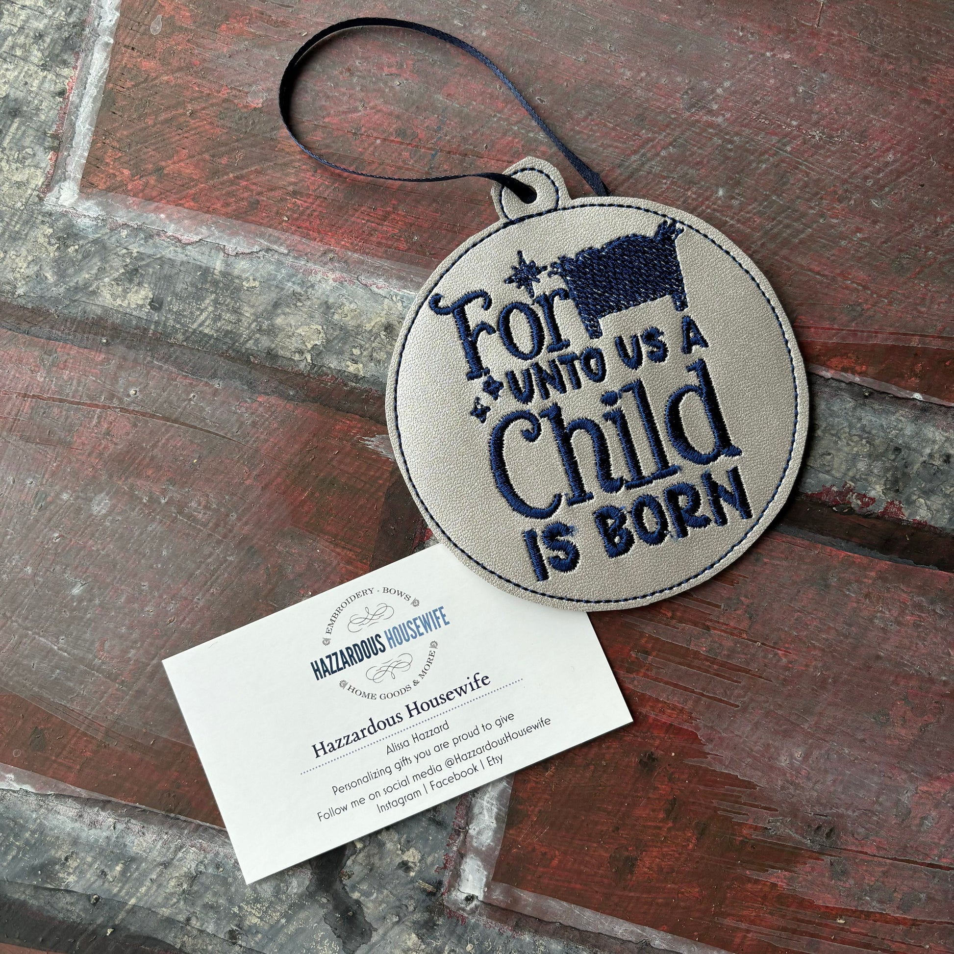 Round embroidered patch with text 'For unto us a child is born' on a wooden surface, accompanied by a business card.
