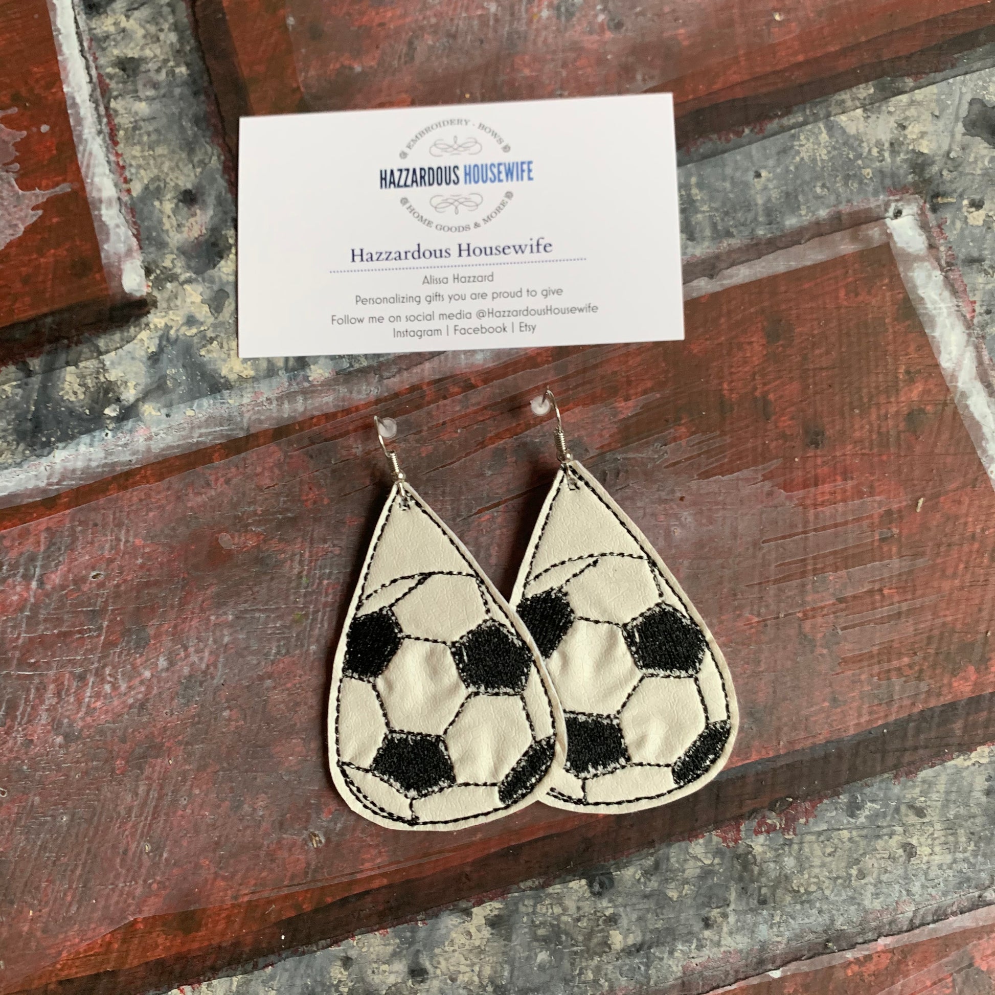 Teardrop-shaped soccer ball patterned earrings on a textured surface with a business card.