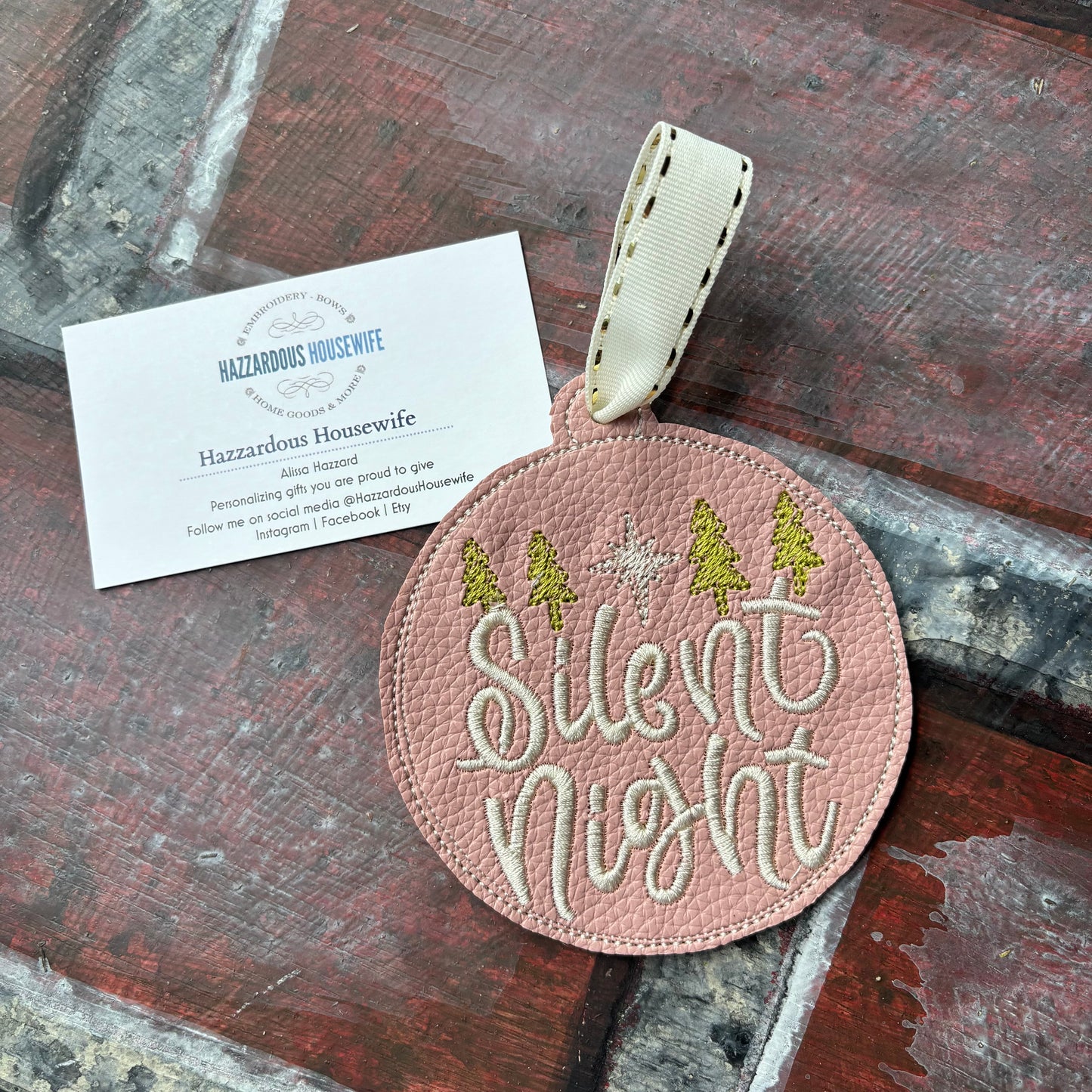 Round pink embroidered tag with 'Silent Night' text on a wooden surface, featuring a HazzardousHousewife business card below.