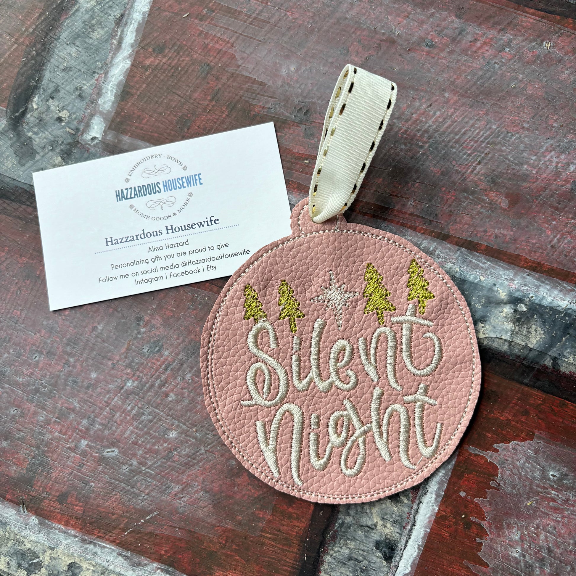 Round pink embroidered tag with 'Silent Night' text on a wooden surface, featuring a HazzardousHousewife business card below.
