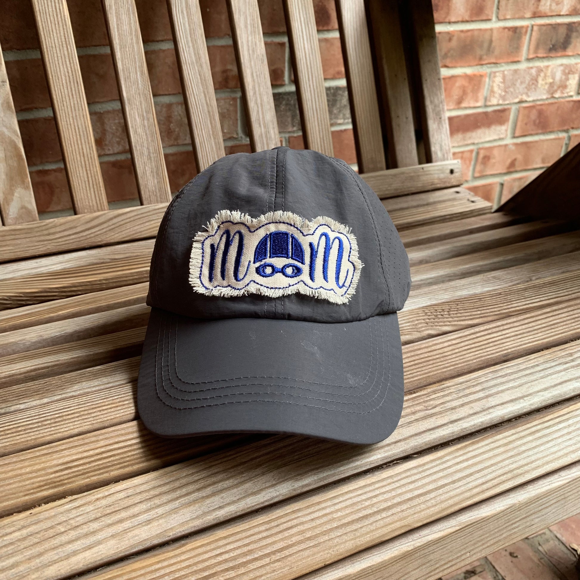 Dark gray cap with 'mom' patch on a wooden bench.  The O in mom is a swim cap and goggles.