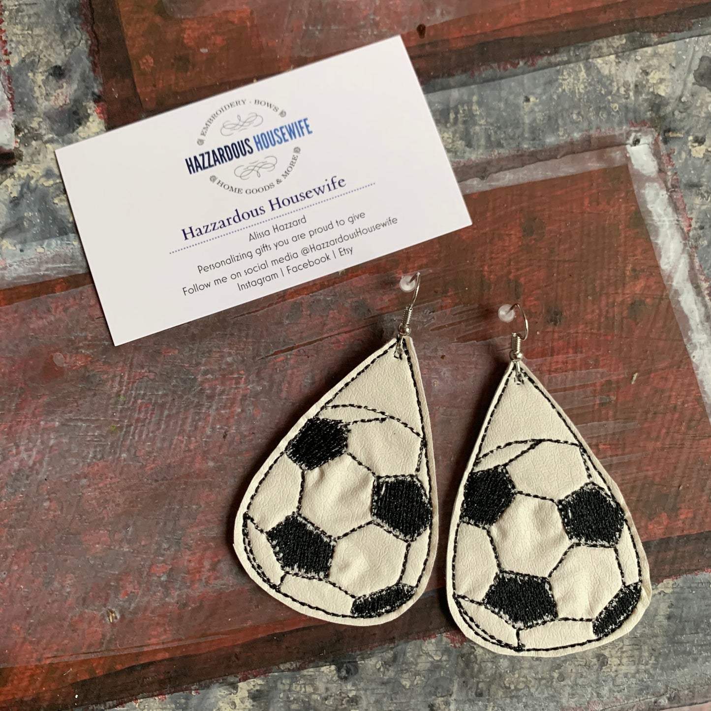 Teardrop-shaped soccer ball earrings on a wooden surface with a business card.