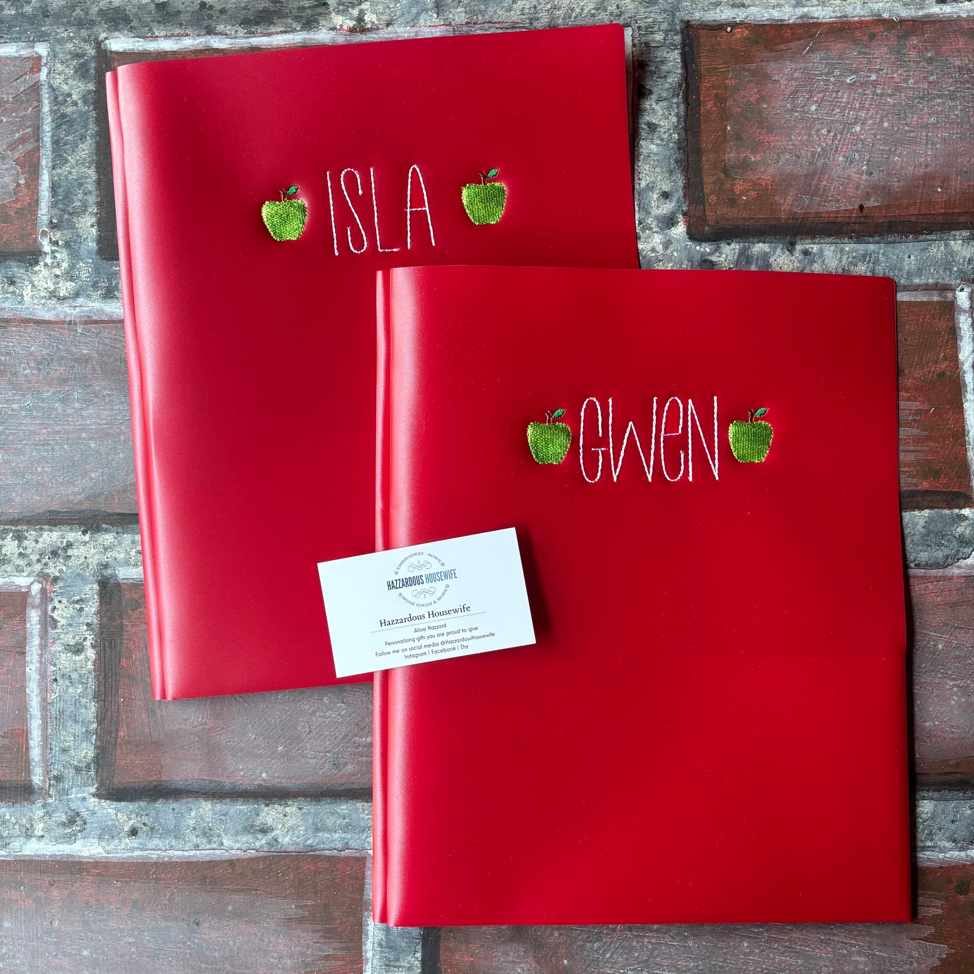 Two red plastic folders with embroidered names and green apples on a brick surface