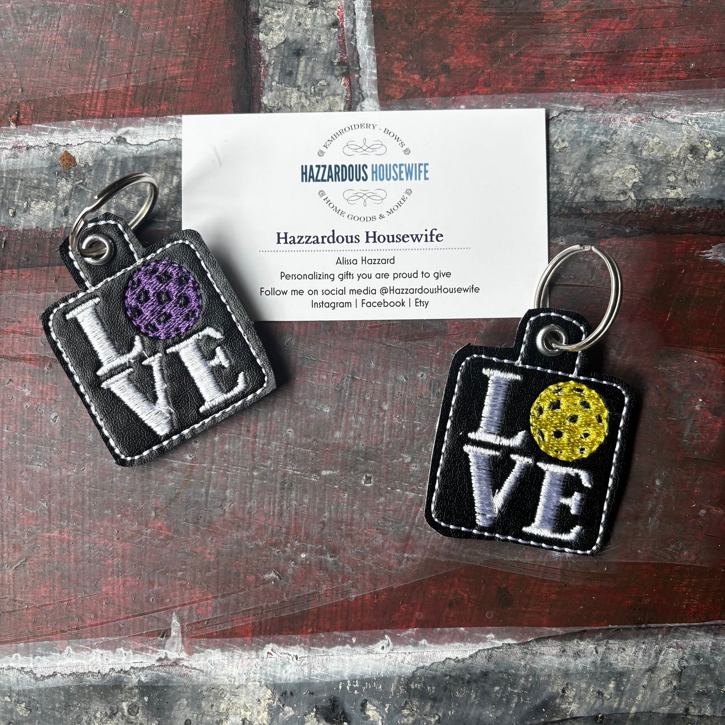 Two pickleball keychains with 'LOVE' text and colorful centers on a rustic surface with a business card.