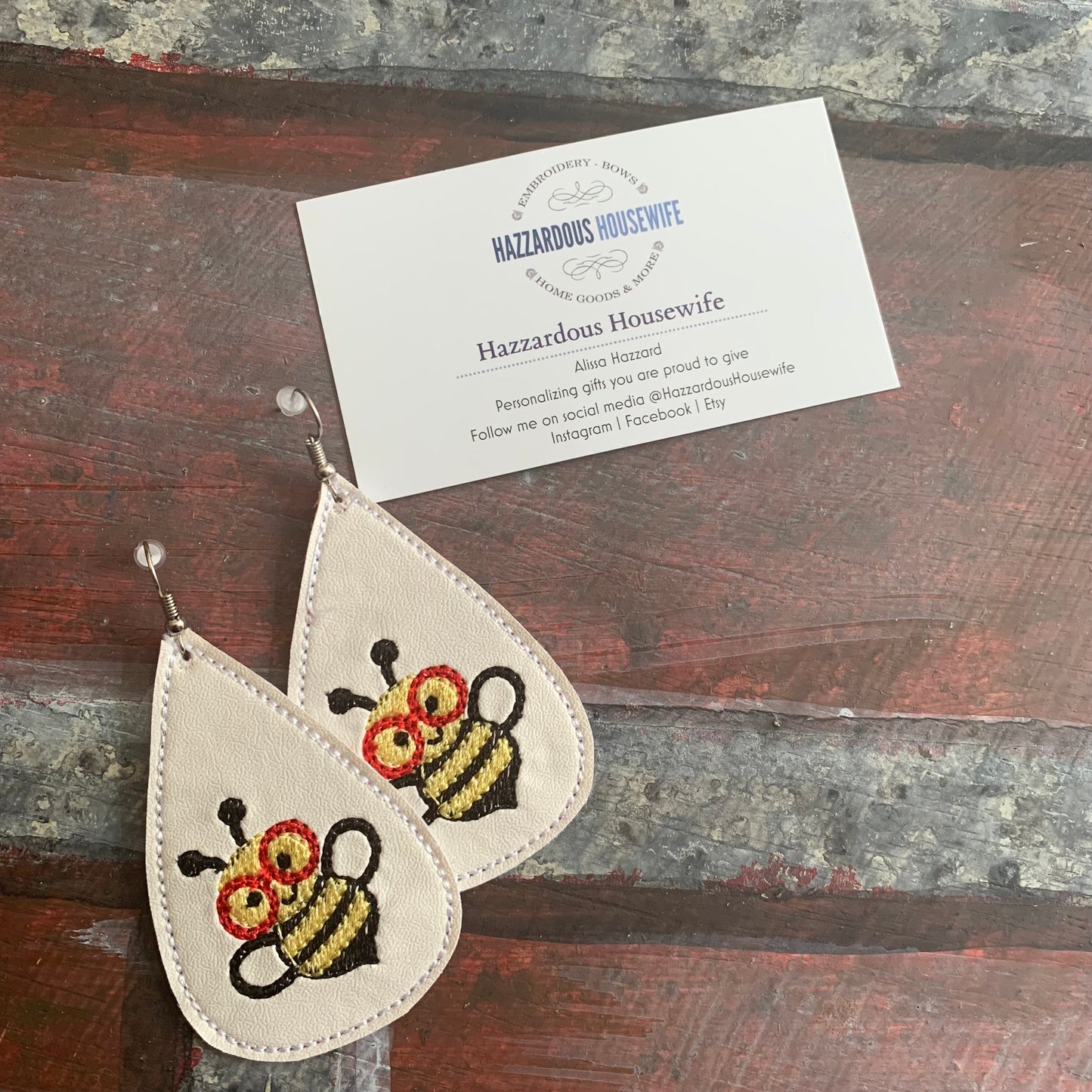 Pair of white teardrop-shaped earrings with bee designs on a wooden surface, accompanied by a business card for 'Hazzardous Housewife'.