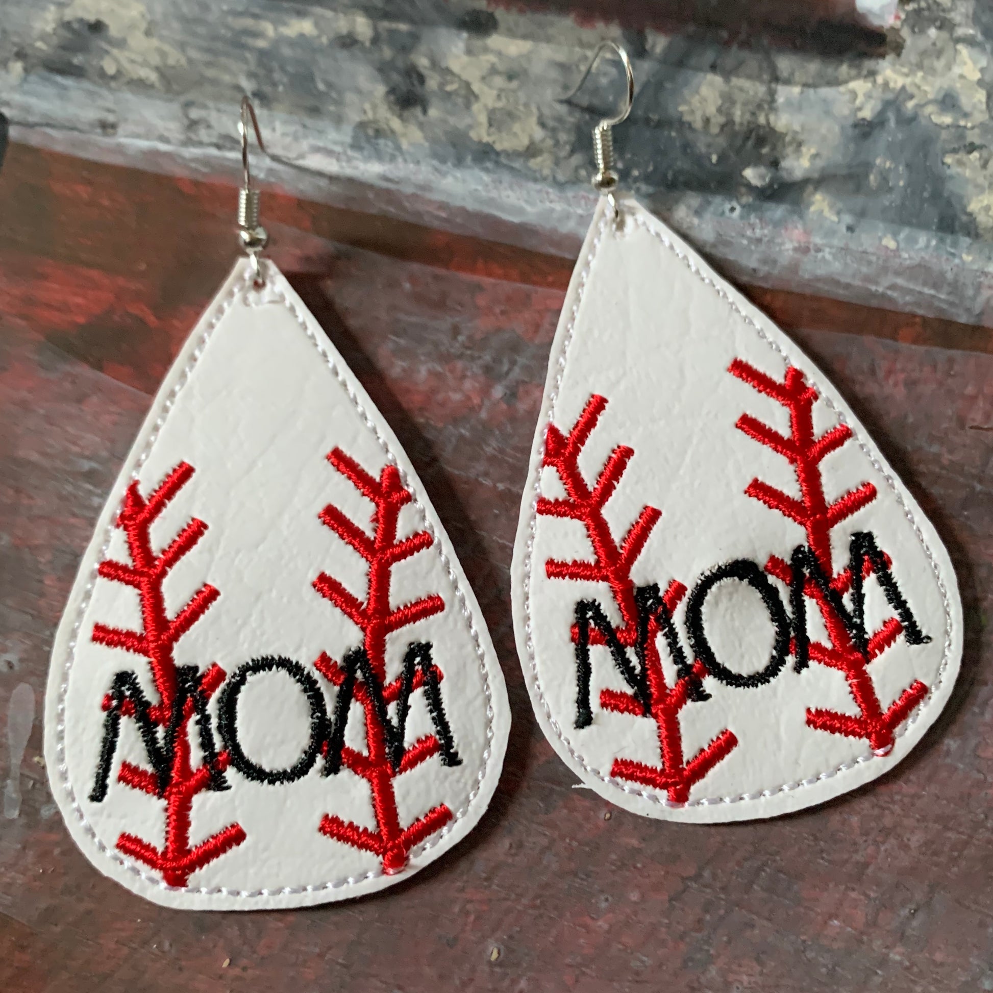 Teardrop-shaped earrings with 'MOM' embroidery on a wooden surface