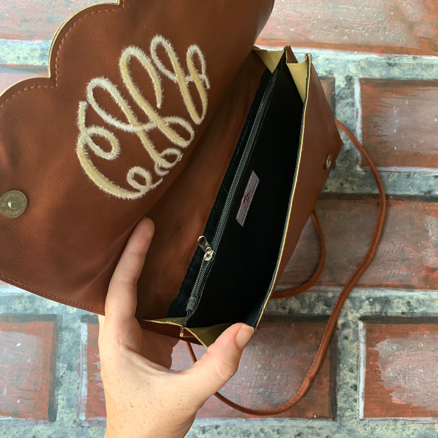 Brown leather bag being held open to show zipper compartment with  monogram on a brick surface