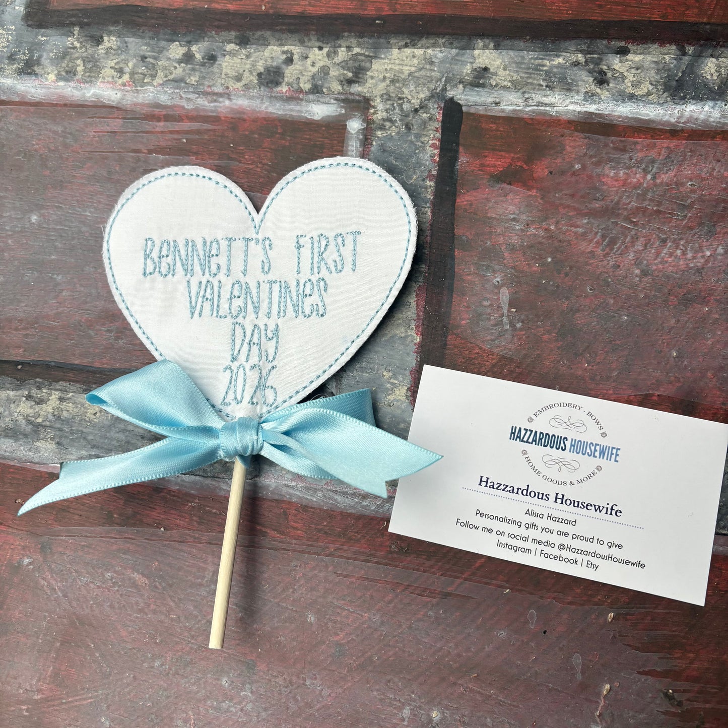 Heart-shaped sign on a stick with 'Bennett's First Valentine's Day 2026' text, tied with a blue ribbon, on a rustic wooden background.