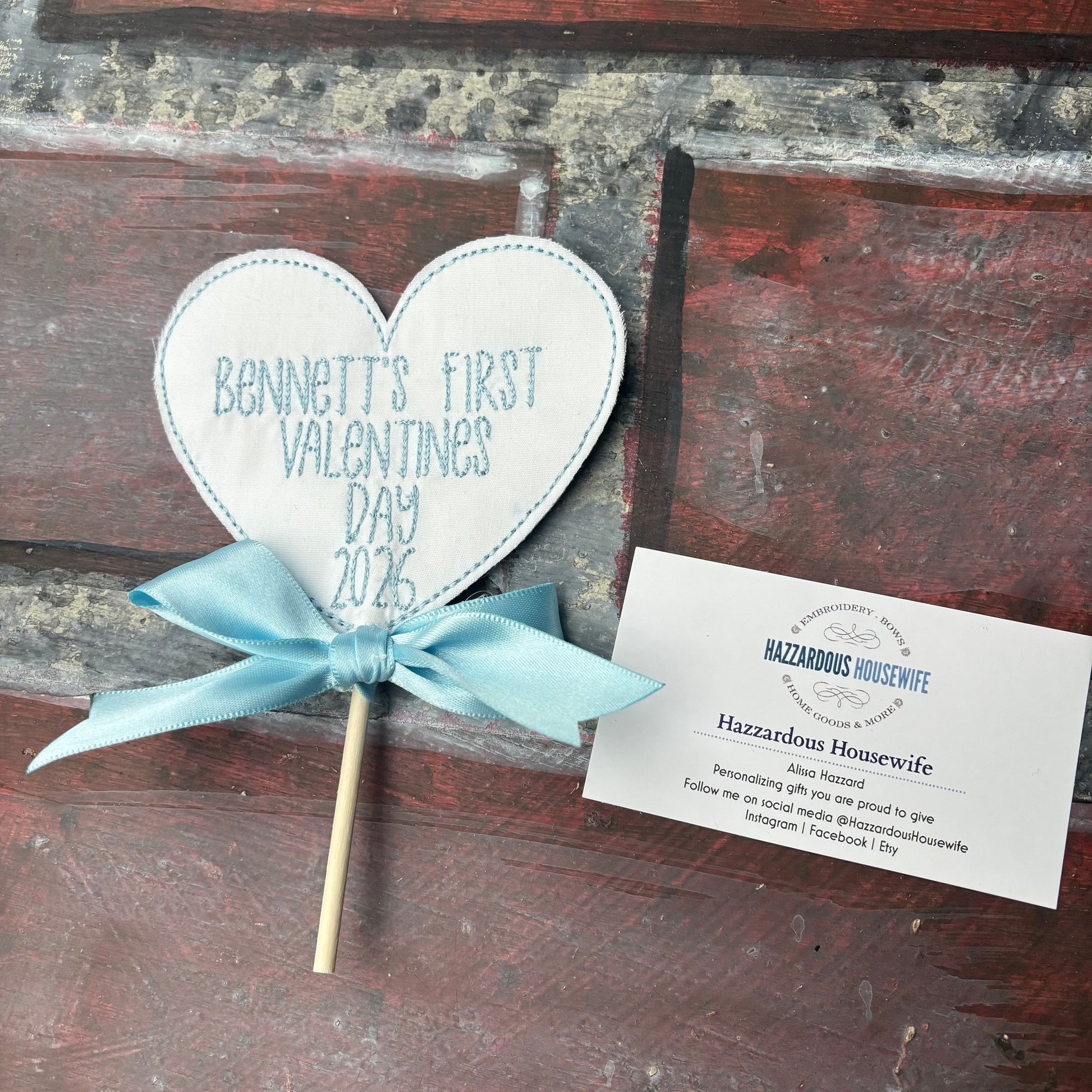 Heart-shaped sign on a stick with 'Bennett's First Valentine's Day 2026' text, tied with a blue ribbon, on a rustic wooden background.