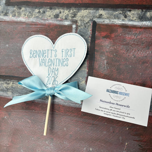 Heart-shaped sign on a stick with 'Bennett's First Valentine's Day 2026' text, tied with a blue ribbon, on a rustic wooden background.