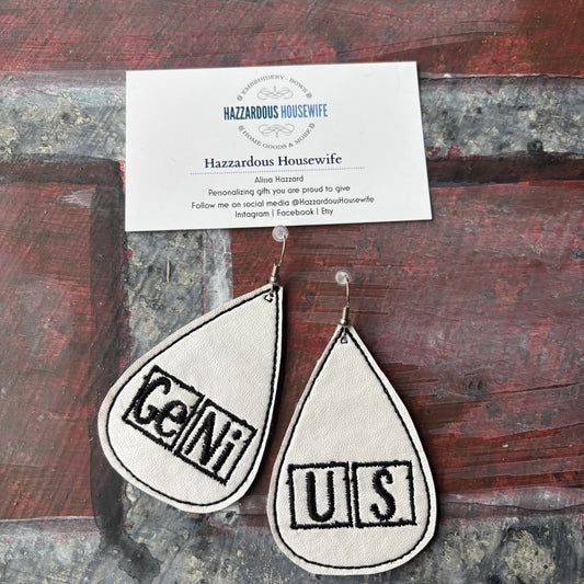 Teardrop-shaped earrings with 'GeNiUS' periodic table text on a wooden background, accompanied by a business card for 'Hazzardous Housewife'.