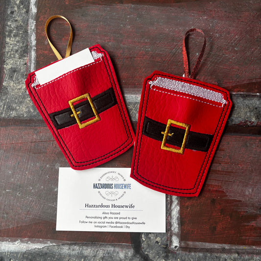 Two red coffee cup-shaped ornaments with a belt buckle to look like Santa's belt on a wooden surface, accompanied by a card from Hazzardous Housewife.